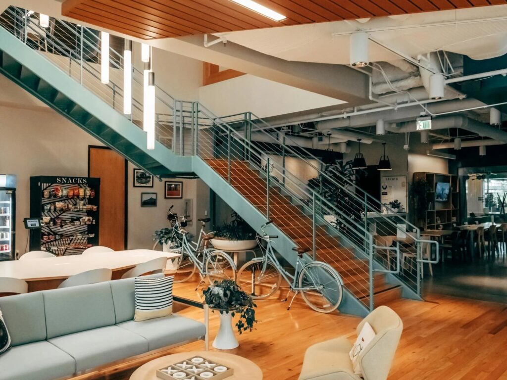 Zone de détente de bureau confortable avec des sièges moelleux, des vélos garés près d'un escalier, et un distributeur de collations en arrière-plan.