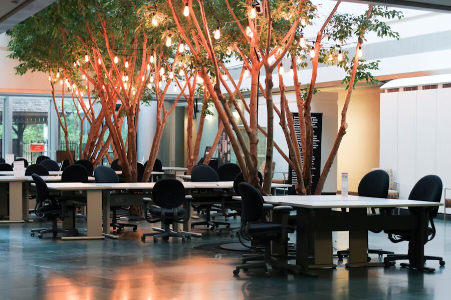 Grand espace de coworking intérieur décoré d’arbres naturels et d’ampoules suspendues, avec de nombreux bureaux partagés et fauteuils de bureau noirs.