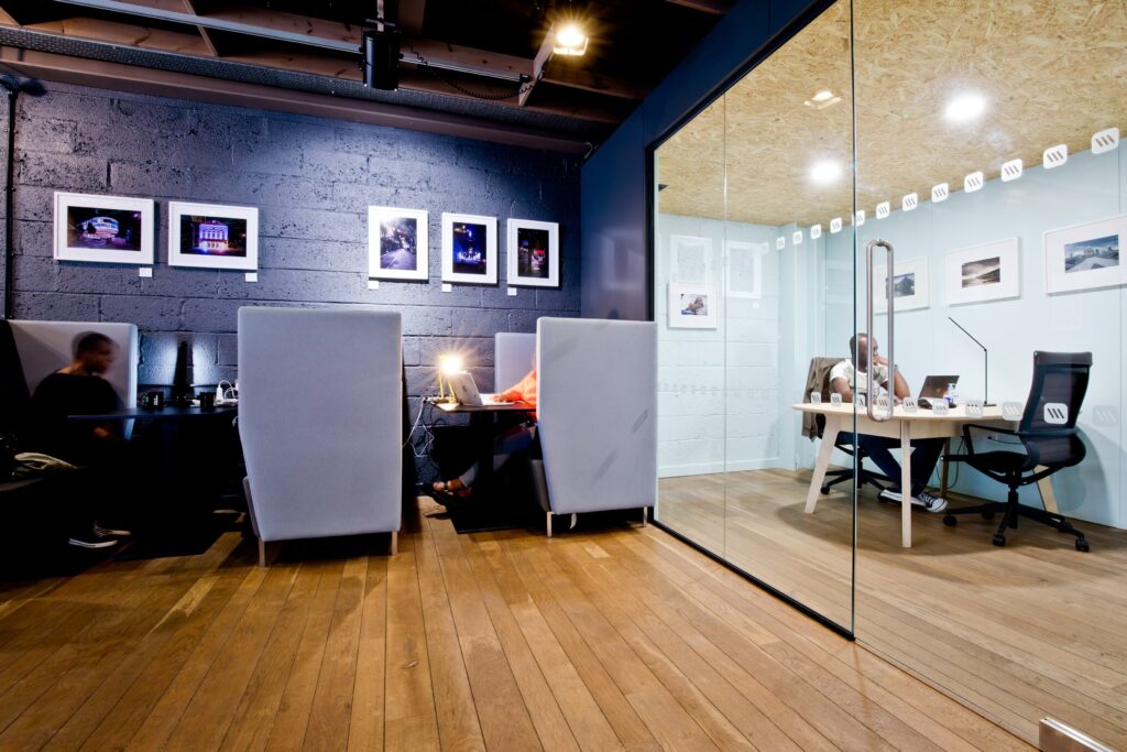 Espace de travail partagé avec alcôves dotées de fauteuils hauts, murs foncés décorés de photos encadrées, et salle vitrée pour réunions privées.